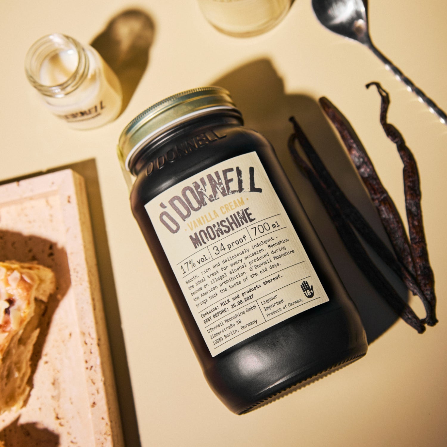 Vanillecrème Moonshine 700 ml pot op crèmekleurige achtergrond naast een shot vanille moonshine en vanillebonenpeulen