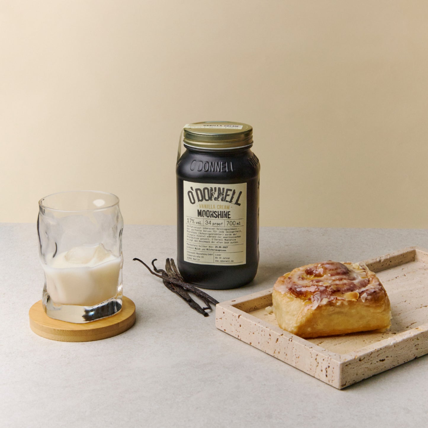 Vanillecrème Moonshine 700 ml pot op een tafel met een kaneelbroodje en een glas gevuld met vanillemoonshine tegen een crèmekleurige achtergrond
