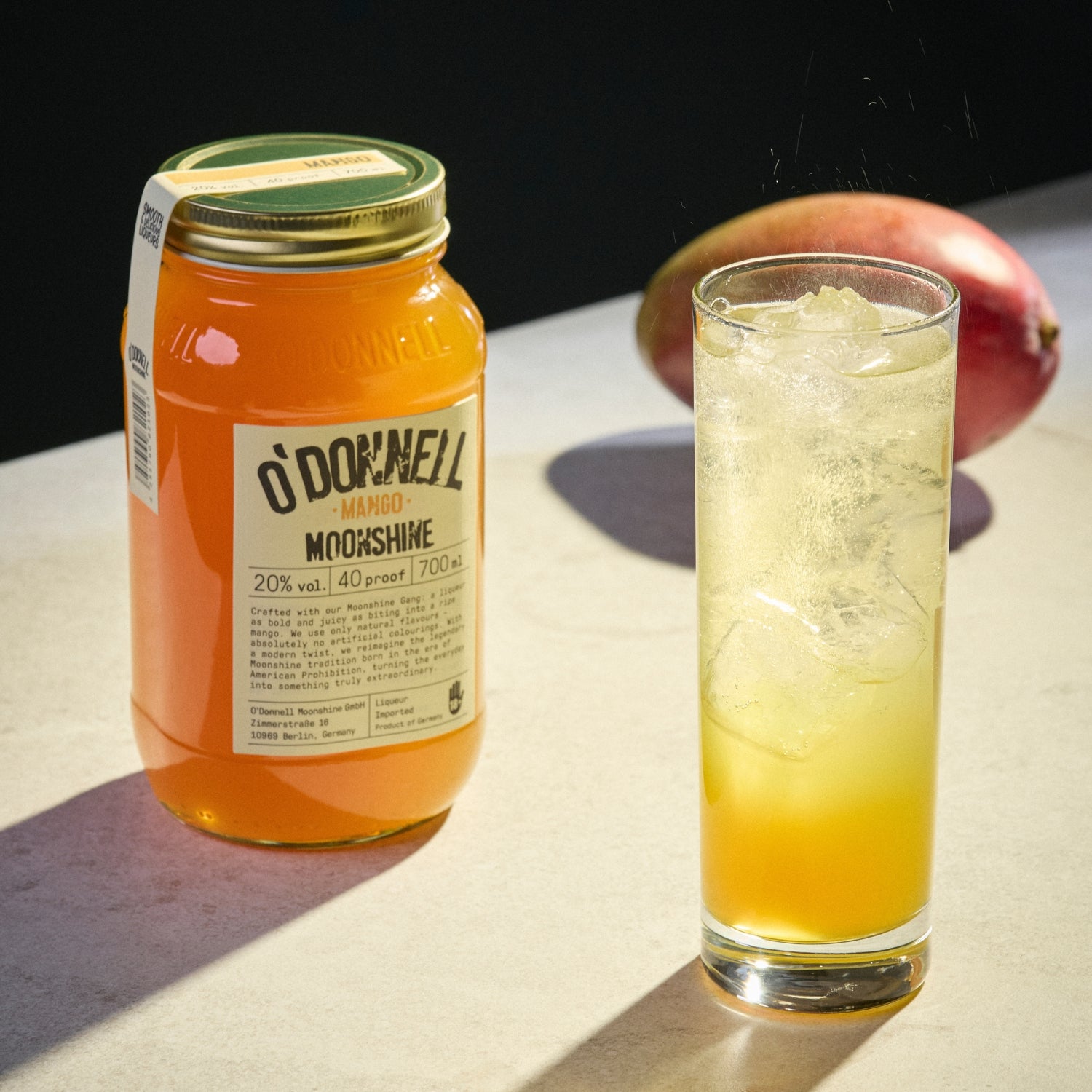 Een mangolikeur aan de linkerkant en een glas gevuld met ijs en de likeur aan de rechterkant, met een echte mango wazig op de achtergrond.