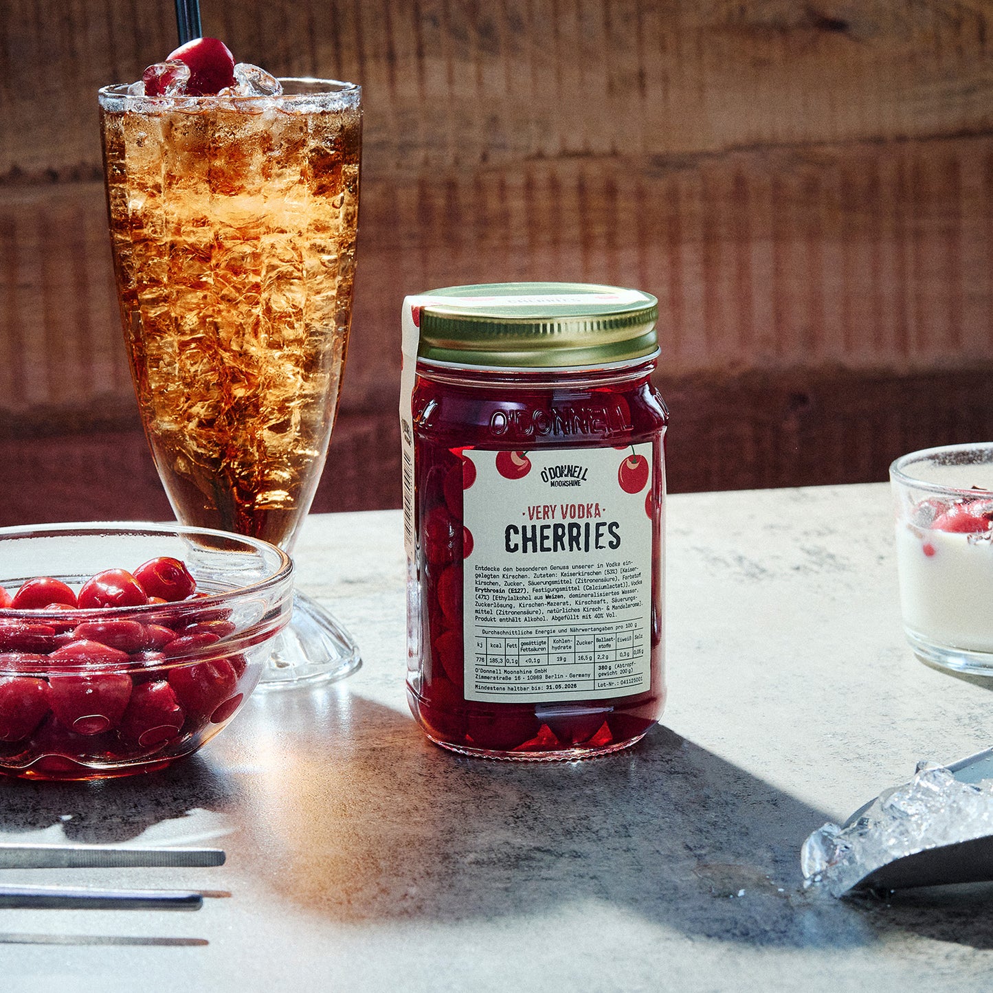 Potje Very Vodka Cherries tegen een houten achtergrond naast een ijsdrankje en een schaaltje kersen
