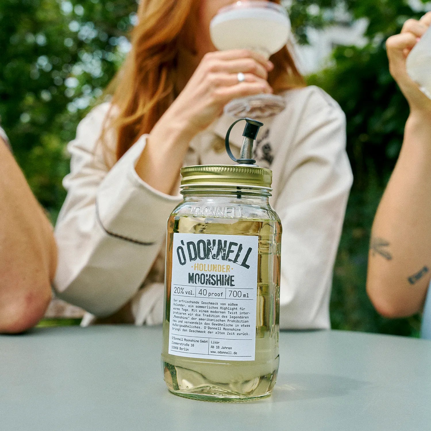 O'Donnell Moonshine vlierbessenlikeur in een Mason Jar, met op de achtergrond een groep mensen die ervan genieten als cocktail.